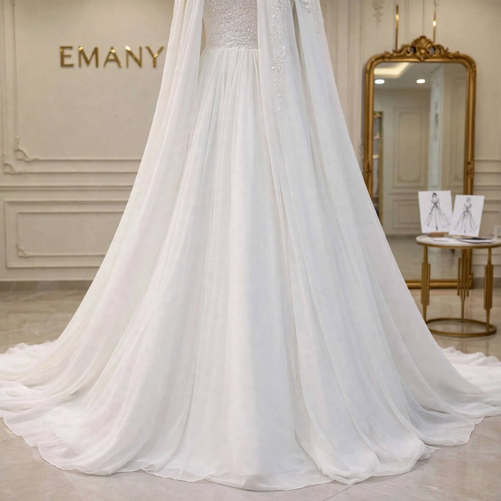 A white bridal gown with a long sheer train and veil displayed indoors on a mannequin or dress form. The background shows a cream-colored wall with the brand name EMANY, a gold-framed mirror, and a small table with sketch cards.