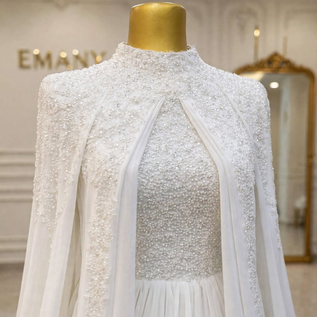 A white embellished high-neck dress displayed on a gold mannequin torso in an elegant indoor setting. The dress features intricate beadwork and sequin detailing, sheer draped panels at the front, and a soft pleated skirt.