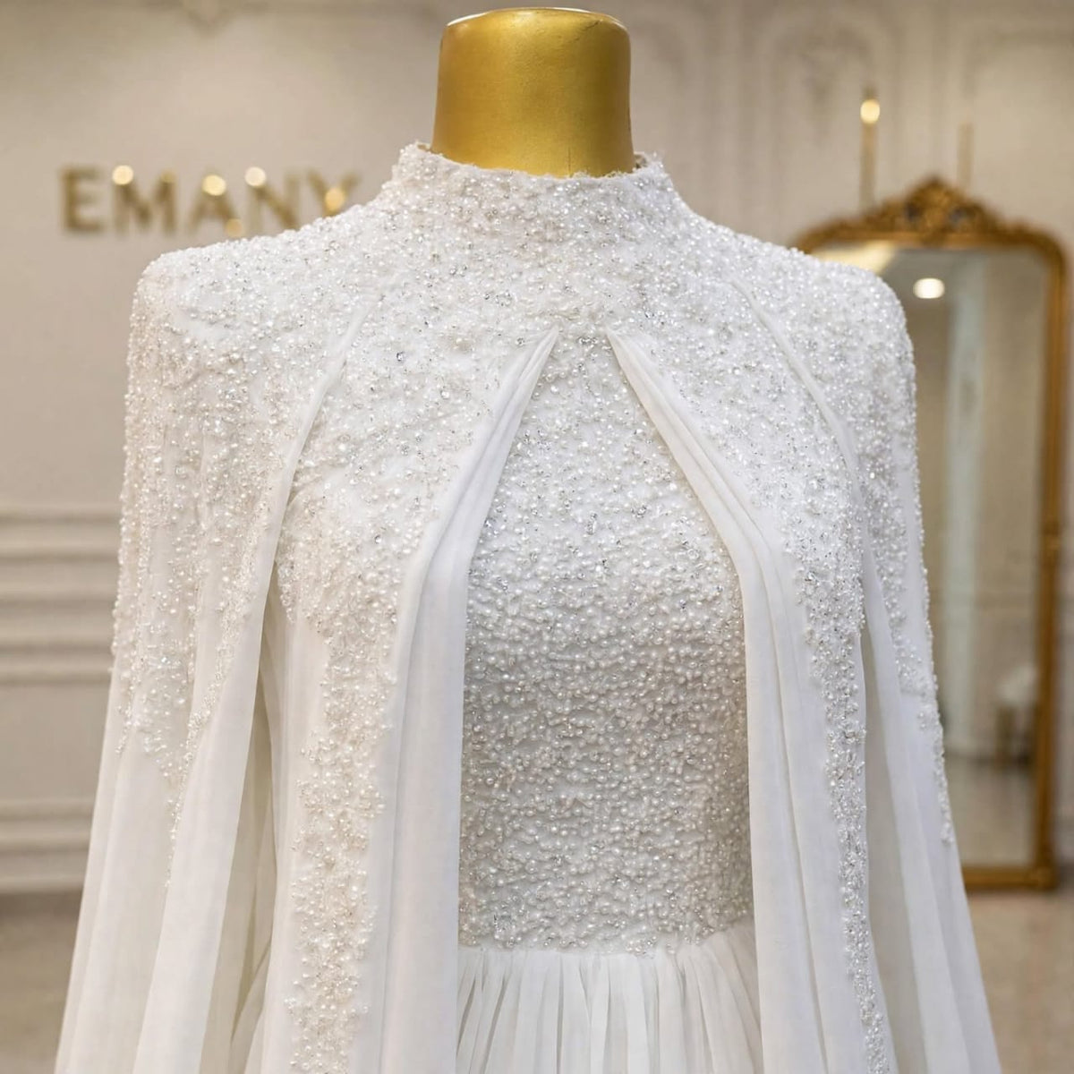 A white embellished high-neck dress displayed on a gold mannequin torso in an elegant indoor setting. The dress features intricate beadwork and sequin detailing, sheer draped panels at the front, and a soft pleated skirt.