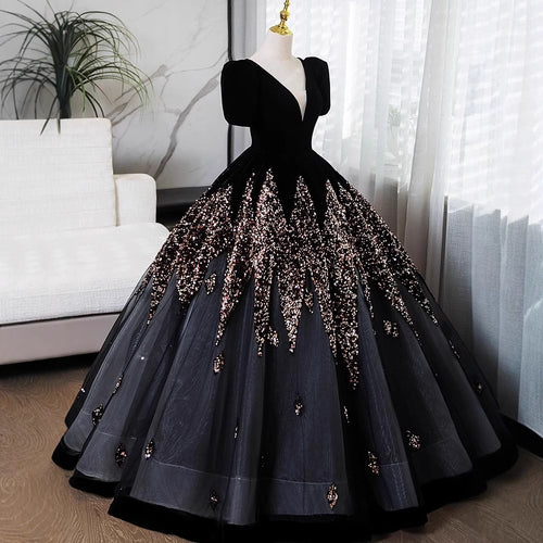 A formal ball gown displayed on a mannequin in an indoor room. The dress has a black velvet bodice with short puff sleeves and a very full dark gray tulle skirt heavily embellished with rose-gold sequins and beadwork.