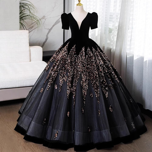A formal ball gown displayed on a mannequin in an indoor room. The dress is black with short puff sleeves, a deep V-neckline, and a voluminous tulle skirt heavily embellished with sparkling sequins and beadwork in a champagne-gold tone.