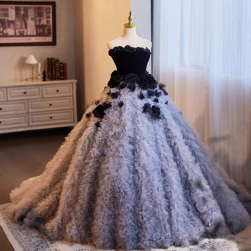 A voluminous ball gown displayed on a dress form in an indoor room. The dress has a black strapless bodice with floral appliqué and a very full skirt made of layered, ruffled fabric in shades of gray and black, with a matching gray train extending behind it.