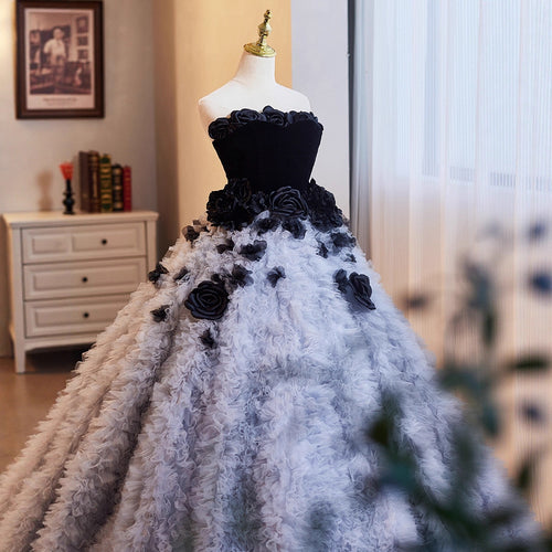 A voluminous ball gown displayed on a dress form mannequin in an indoor room. The dress has a black strapless bodice with 3D floral appliqués and a matching black floral waistband, transitioning into a very full skirt made of textured, feather-like or ruffled white fabric with scattered black floral accents.