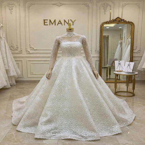 A white ball gown wedding dress with a sweetheart neckline, sheer long sleeves, and a heavily embellished sequined skirt, displayed on a mannequin in a bridal boutique with the brand name Emany on the wall.