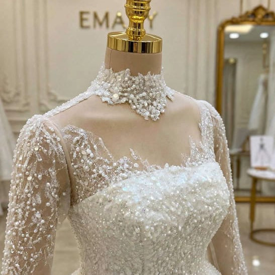 An elegant white wedding gown with long sheer sleeves and dense beadwork and sequin embellishments, displayed on a mannequin in a bridal boutique.