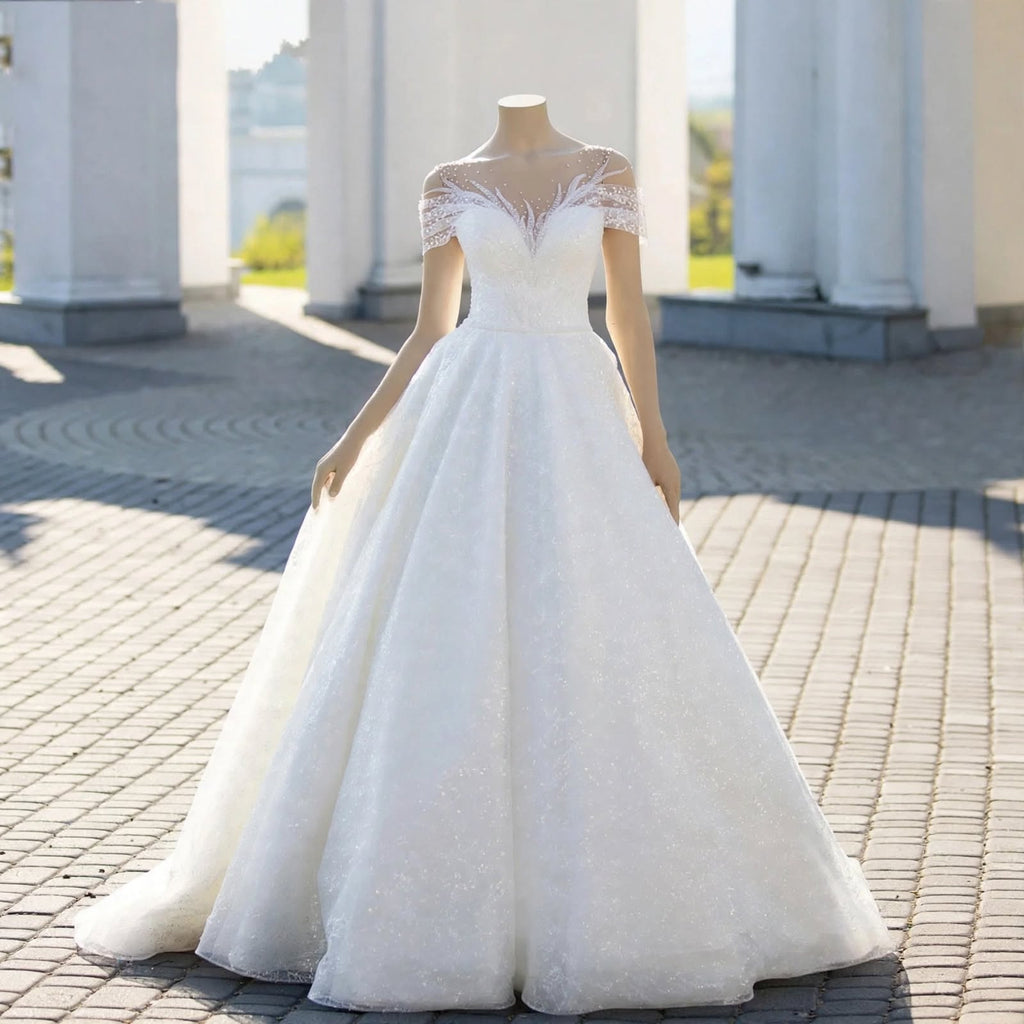 White A-line wedding dress with sheer illusion neckline, lace appliqué, pearl beading, and off-the-shoulder sleeves displayed on a mannequin.

