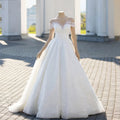 White A-line wedding dress with sheer illusion neckline, lace appliqué, pearl beading, and off-the-shoulder sleeves displayed on a mannequin.

