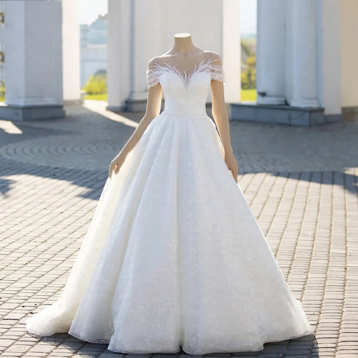 White A-line wedding dress with sheer illusion neckline, lace appliqué, pearl beading, and off-the-shoulder sleeves displayed on a mannequin.


