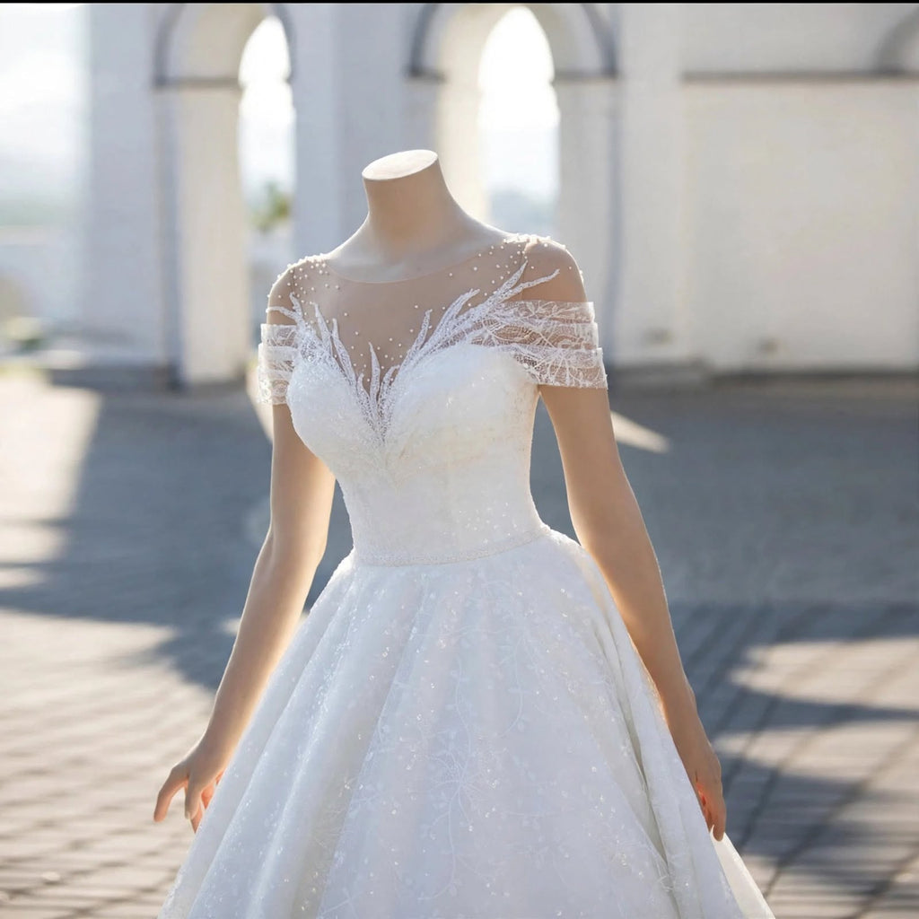 White A-line wedding dress with sheer illusion neckline, lace appliqué, pearl beading, and off-the-shoulder sleeves displayed on a mannequin.


