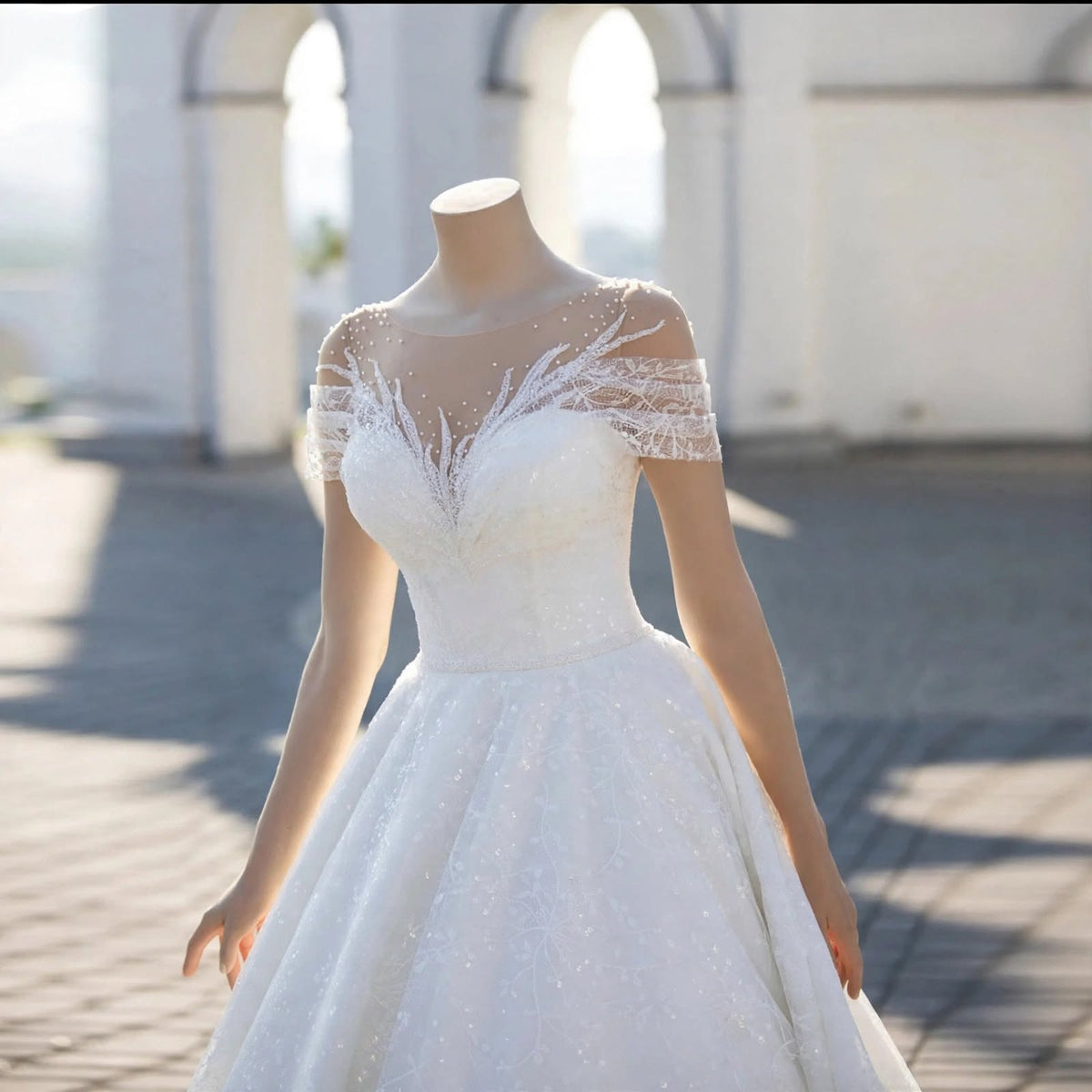 White A-line wedding dress with sheer illusion neckline, lace appliqué, pearl beading, and off-the-shoulder sleeves displayed on a mannequin.


