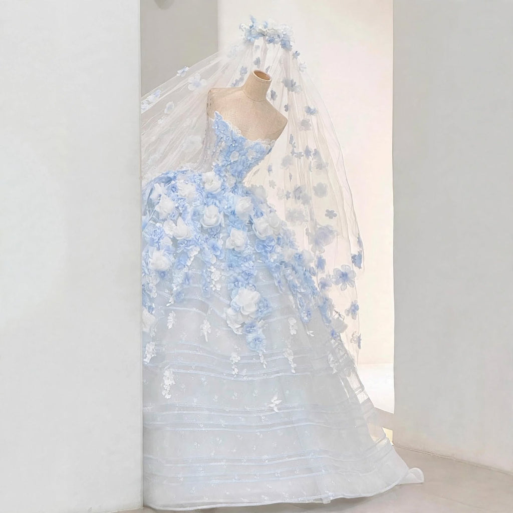 Light blue ball gown wedding dress with white and blue 3D floral appliqués, sheer corset bodice, and a matching floral cathedral veil displayed on a mannequin.

