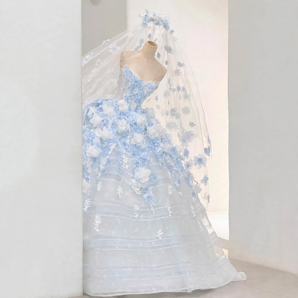Light blue ball gown wedding dress with white and blue 3D floral appliqués, sheer corset bodice, and a matching floral cathedral veil displayed on a mannequin.

