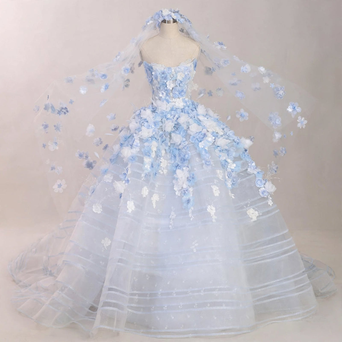 Light blue ball gown wedding dress with white and blue 3D floral appliqués, sheer corset bodice, and a matching floral cathedral veil displayed on a mannequin.

