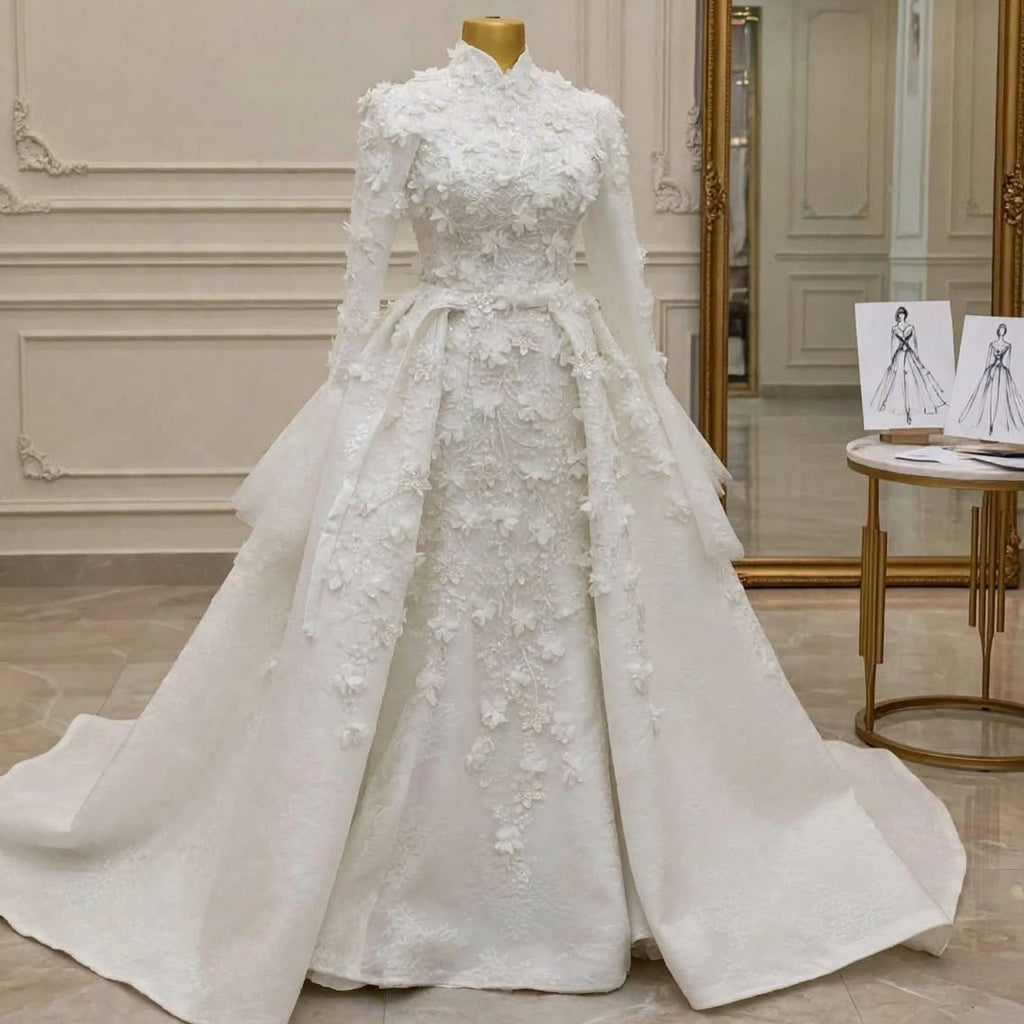 A white long-sleeve wedding gown with intricate floral embroidery and a high collar, displayed on a mannequin in a luxury bridal boutique with the brand name EMANY on the wall.