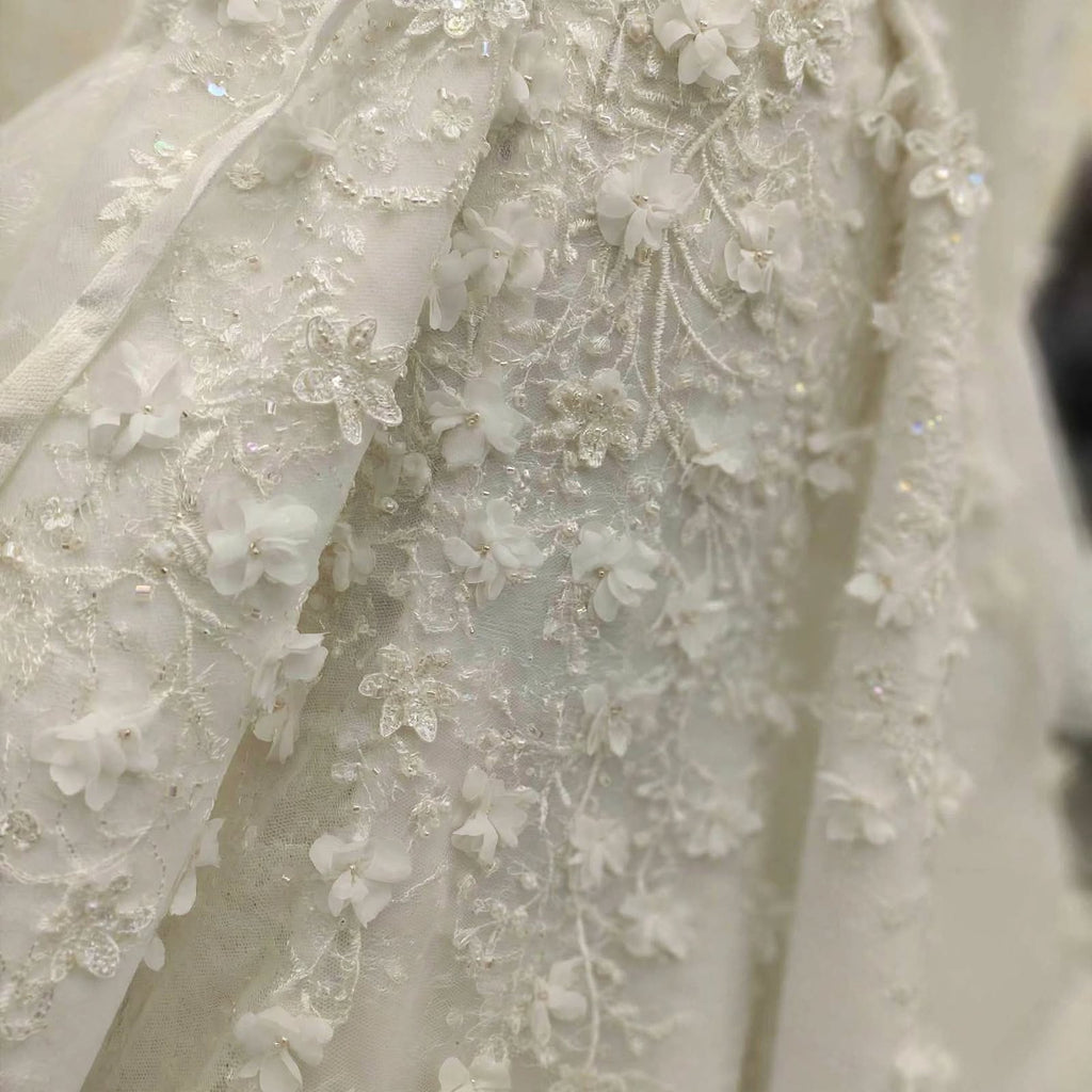 Close-up of a white bridal gown fabric featuring intricate floral lace applique, beadwork, and 3D flower embroidery.
