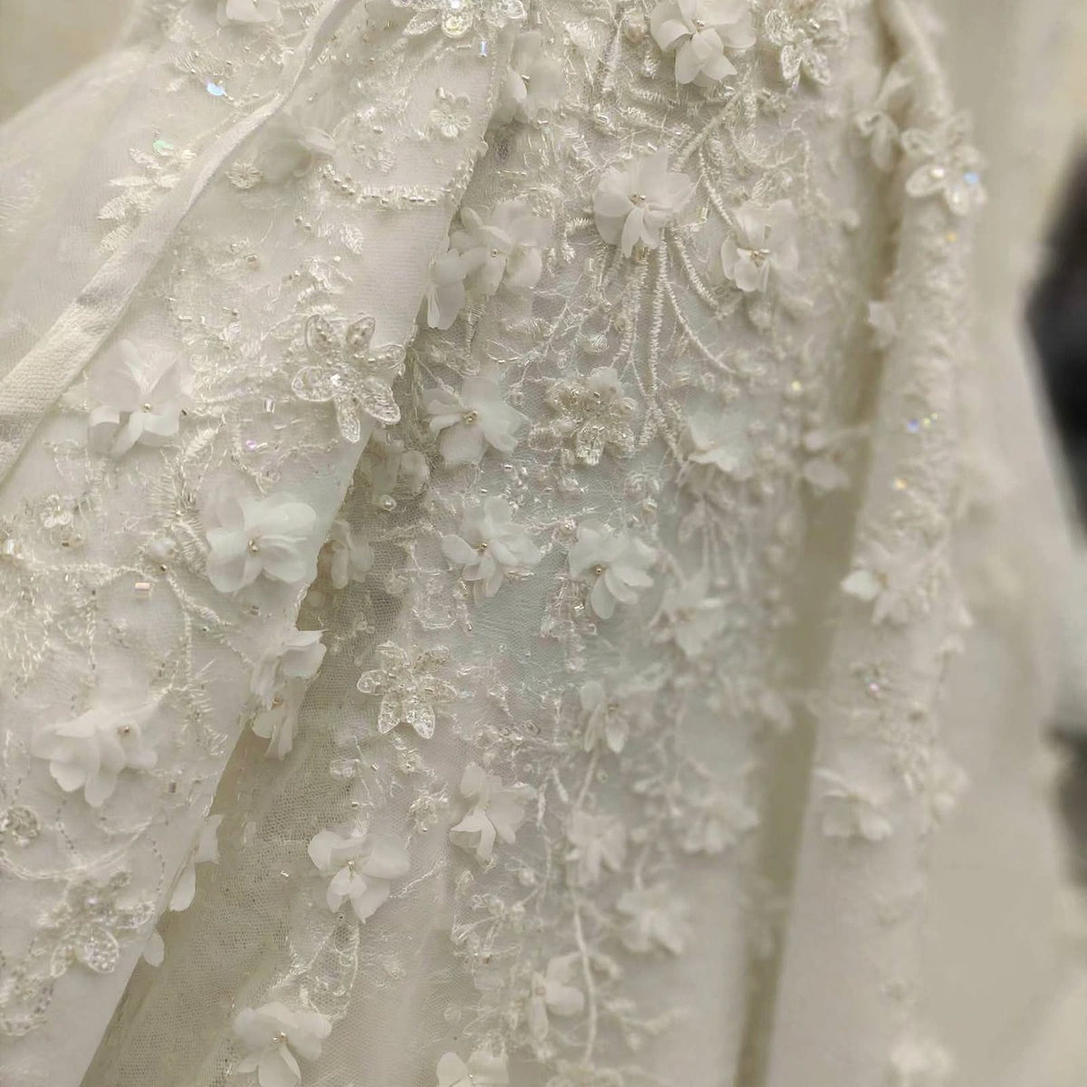 Close-up of a white bridal gown fabric featuring intricate floral lace applique, beadwork, and 3D flower embroidery.
