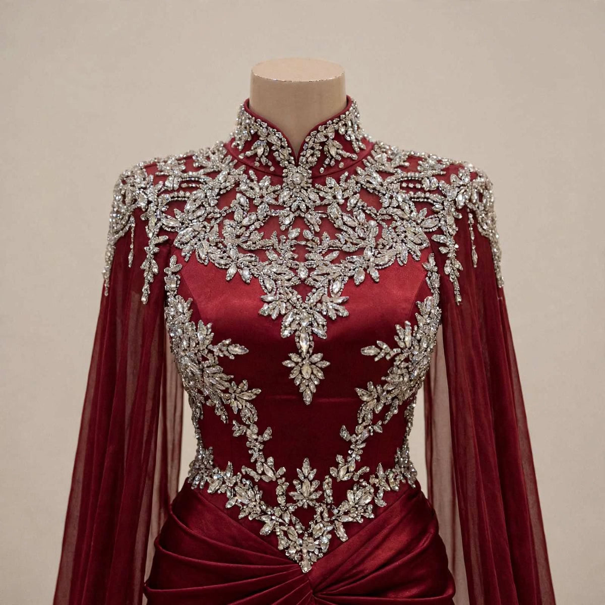 A high-neck, long-sleeved burgundy gown displayed on a mannequin against a plain beige background. The dress features intricate silver beaded floral embroidery across the bodice, shoulders, and neckline, with sheer tulle sleeves and a draped waist detail.