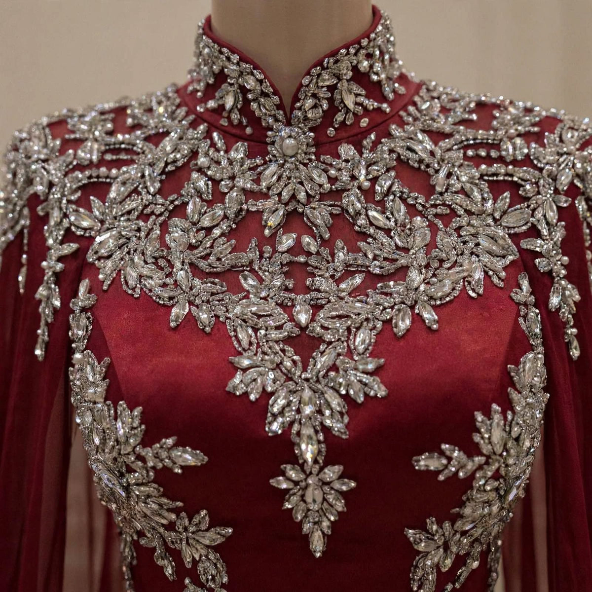 A close-up of a high-neck, long-sleeved red dress or gown on a mannequin, heavily decorated with intricate silver beading and crystal embellishments across the bodice and shoulders.
