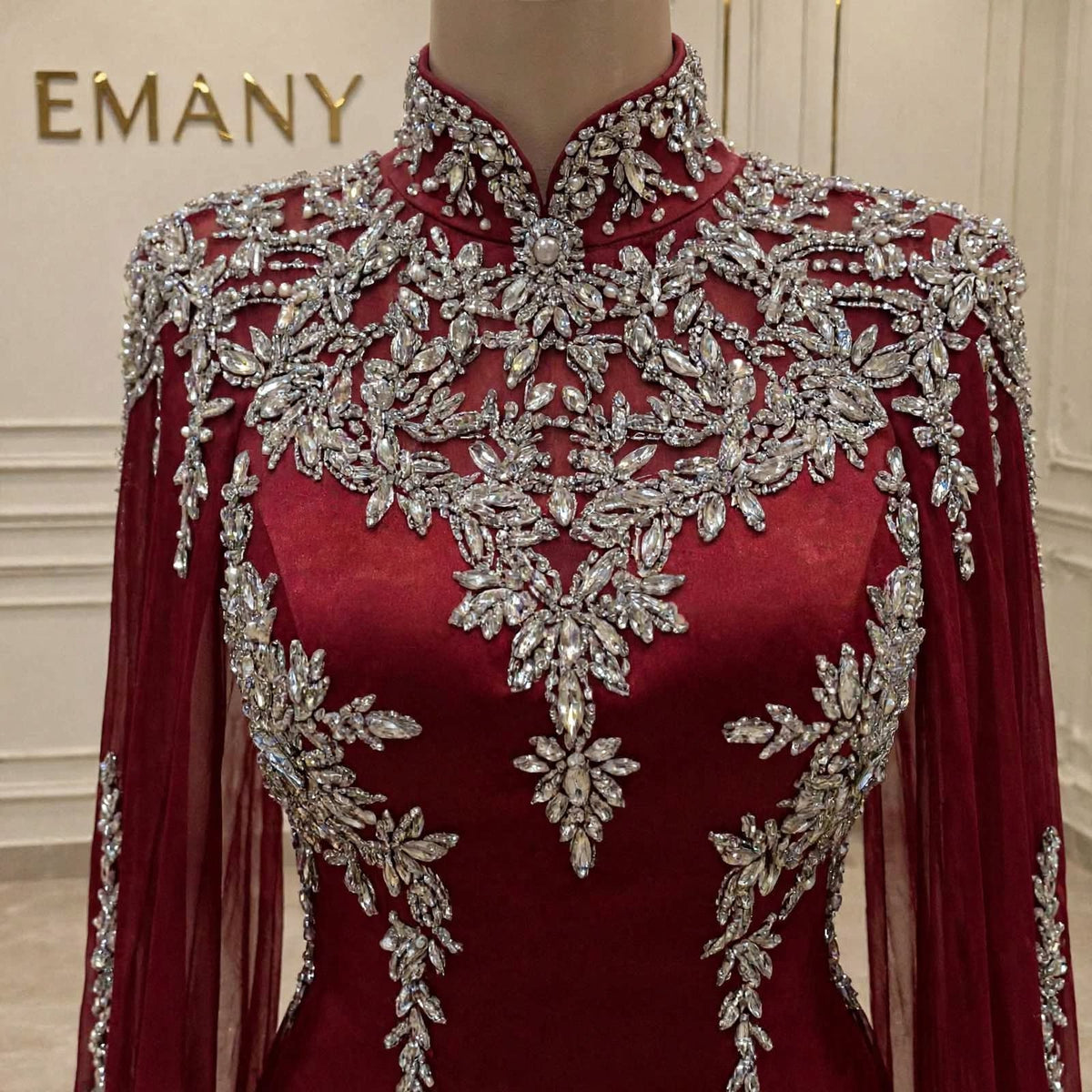 A close-up of a high-neck, long-sleeved burgundy dress displayed on a mannequin. The dress features extensive silver beaded floral embroidery across the bodice, shoulders, and sleeves.